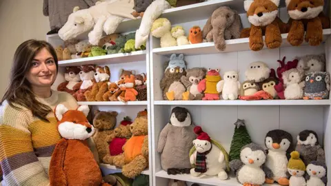 Amanda Hope Amanda Hope stands holding a toy fox next to her collection of Jellycat toys stacked on shelves