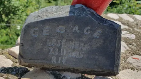 Salvamont Brașov A smooth dark rock which says "George, lost here, 23.11.2025, RIP". It has been placed on a mound of other rocks.