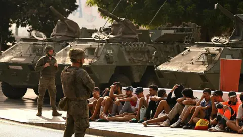 AFP via Getty Images Military personnel stand near armored vehicles while a group of seated people wait on a curb beside a road, under trees in daylight