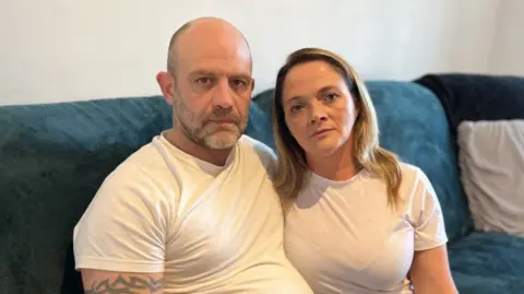 Adam and Kelly Parker sitting on a blue sofa with their arms around each other, wearing white t-shirts and looking straight to camera with blank faces