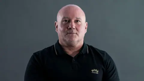 Bedfordshire Police Darren Attwood wearing a black polo shirt with gold detailing on it. He has a shaved head and looks serious.
