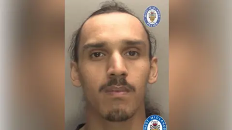 West Midlands Police A man with tied-back dark hair and a trimmed beard and moustache looks at the camera against a grey background in a police mugshot with West Midlands Police logos on it.