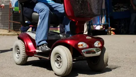 A person sits on a motorised four-wheel, red scooter. They have black trainers on and blue trousers.