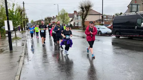 A group of people are running on a wet street during a race, with houses and parked vehicles along the road.