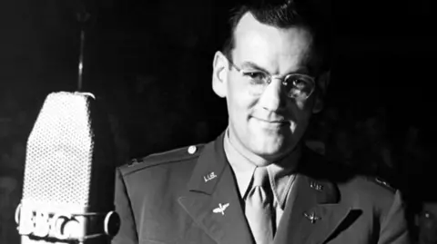 A black and white image of Glenn Miller taken in 1944, he is standing next to a microphone that says BBC. He is smiling to the camera, is wearing a uniform and glasses. He has slicked back hair. 