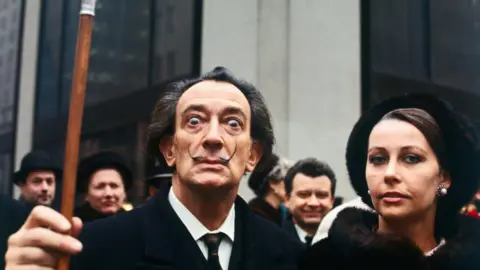 Getty Images The artist Salvador Dali, wearing a dark suit and tie and white shirt, stares into the camera and lifts his walking cane with his right hand. He is standing next to a woman and in front of a large group of people, all wearing black.