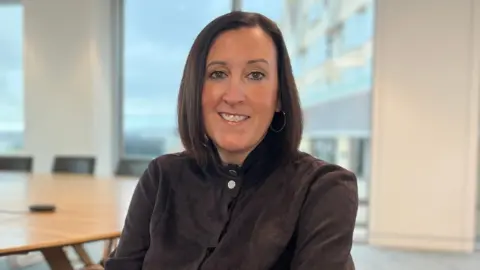 Alex sits in a modern conference room with large windows overlooking a cloudy waterfront. She is wearing a dark long‑sleeved dress and is seated on a chair near a long wooden meeting table.