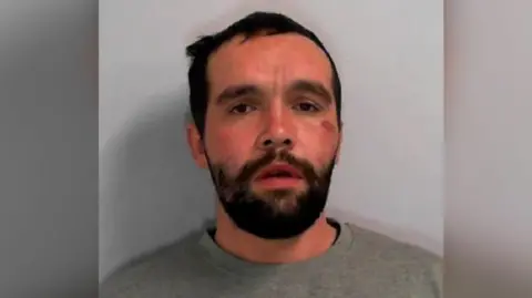 North Yorkshire Police Custody picture of Liam James Watson, 32, a dark haired man with a beard, wearing a grey sweatshirt.