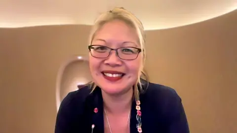 A women with blonde hair and glasses smiles at the camera with a meditiation room in the background.
