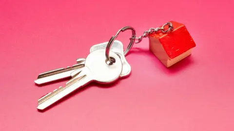 A set of house keys with a house shaped fob on a pink surface.