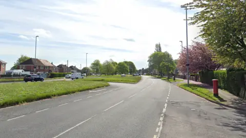 A semi urban street main road lined with trees and semi-detatched houses