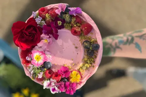 KIERSTEN GEORGE A round cake with pink icing, with flowers and fruit decorated on the circumference of the cake.