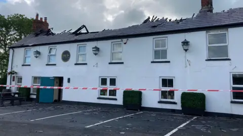 A cordon is set up around a two-storey white building. The roof has been fire damaged and has broken apart. A large blue door on the bottom floor is open and the parking spaces in front of the building are empty.