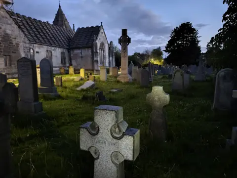 Kevin O'Kane Graveyard at dusk with rows of headstones and a stone church in the background. A tall Celtic cross monument stands among the graves, with soft lighting from nearby buildings under a cloudy evening sky