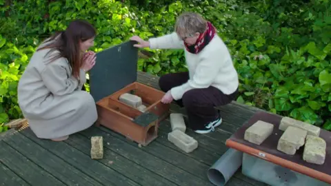 Two women kneeling by a wooden hedgehog house on a deck, surrounded by greenery and loose bricks.