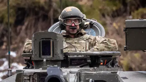 Prince William, wearing a helmet and military camouflage, sitting in a Challenger 2 battle tank