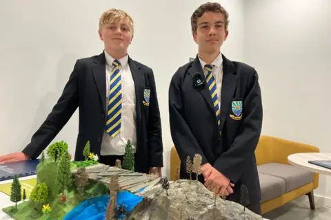 Jon Wright/BBC Two boys in school uniform stand behind a model of an island with a wooden bridge.