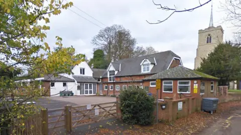 Martin Heath/BBC A primary school with a yard in to the left and old two-storey brick buildings to the right. There is a white building with a triangular upper floor in the background, and a brick wall in the foreground with a wooden gate across the entrance. There is a church to the right.
