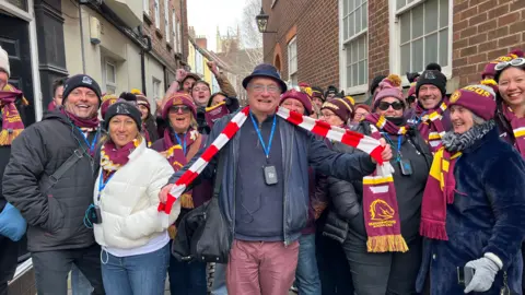 About 20-30 people in red and yellow scarves and hats posing in a historic street smiling at the camera