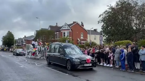 The funeral procession of Elsie Dot Stancombe passes through Birkdale on its way to the church. A hearse pulls ahead of a white coach drawn by white horses, while hundreds of people line the street. 