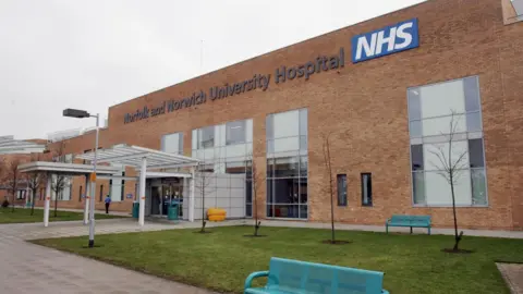 PA Media The entrance to the Norfolk and Norwich University Hospital. It is a light brown brick building with a white entrance canopy. Two turquoise benches are located by a patch of grass.