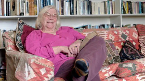 Pacemaker Shows Jennifer Johnston in a pink top with glasses sitting on a patterned sofa with her legs folded and a book on her left knee