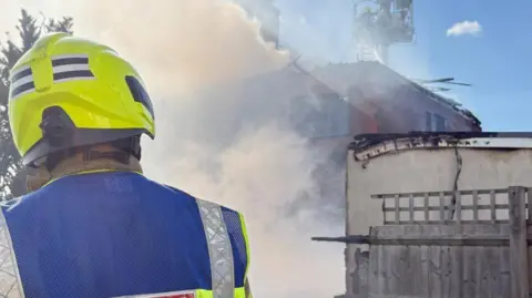 A fire fighter wearing a yellow fluorescent helmet and blue jacket. In the background is smoke coming from a fire at two semi-detached houses.