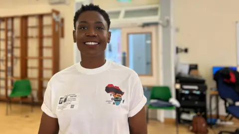 Lesley simles at the camera as she wears a white T-shirt with the charity logo on it. She has short black hair and is standing in a school's sports hall.