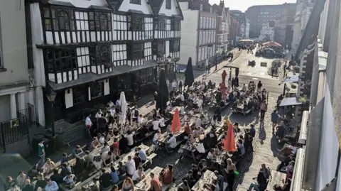 Billie Mallard King Street in central Bristol is visible in a photograph taken from a balcony overhead. Dozens of people are sitting at wooden tables in the street and in the background the dark wood and white paint frontage of the Llandoger Trow pub is visible.