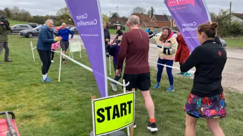 A group of runners setting off on a charity fun run in a park on a cloudy day. Many are wearing sports wear and they are being cheered on by spectators. There are houses in the background.