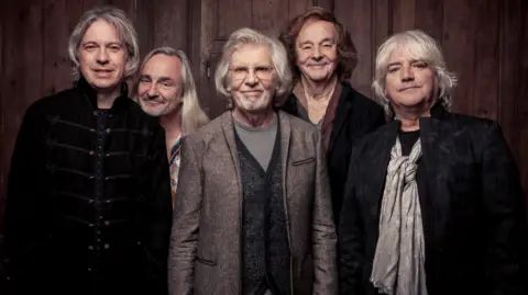 The Zombies band in a recent photo. Rod Argent is centre wearing glasses and a speckled suit jacket and striped T-shirt, he is smiling at the camera and has grey medium-length hair and stubble. 