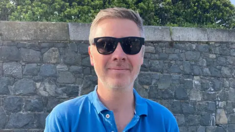 Macey Turner/ BBC Paul Cunningham is wearing a blue shirt and dark glasses. he has some grey stubble on his chin and very short cropped hair.