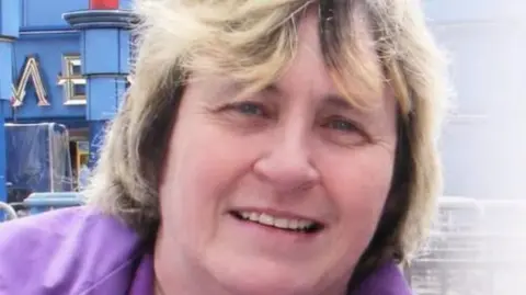 A close up of Theresa Wheelhouse. She has shoulder length blonde/brown hair and is wearing a purple jacket.