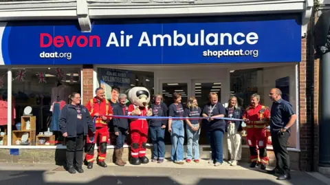 A ribbon-cutting ceremony in front of a store a newly opened Devon Air Ambulance charity shop. There are 11 people stood outside the shop all wearing company uniform. One is dressed as a mascot. 