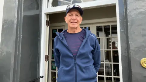 Older man with cap and blue tracksuit top pictured outside hotel entrance.