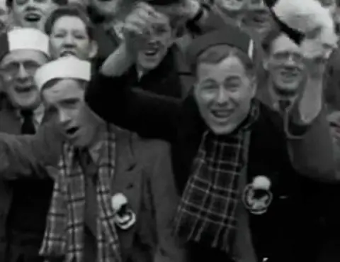 A tightly packed group of spectators fills the frame, gathered closely together in a stadium stand. Several individuals in the foreground wear scarves and hats, with raised arms and open mouths suggesting active cheering or chanting.
The black‑and‑white image captures the dense crowd and energetic match‑day atmosphere.