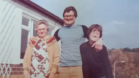 Kenneth S Farrington Kenneth is in the middle of the photograph with his arm around his mum on his right and his son Alan on his left. Alan has his eyes closed in the picture and is smiling. Alan is wearing a dark long sleeve top and has brown hair. Margaret is smiling. She has grey hair and is wearing a mustard cardigan. Kenneth is wearing glasses, a sky blue top with dark sleeves and mustard trousers. 
