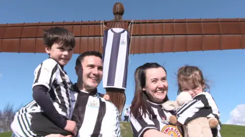 A man and woman wearing Newcastle Unite tops and they are holding two children. The Angel can be seen in the background with a black and white shirt.