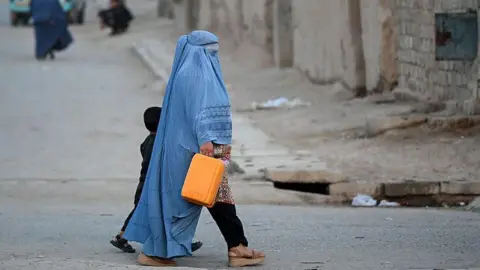 An Afghan burqa-clad woman walks along a street in the 7th district of Afghanistan's Kandahar, several people are behind her. She is walking next to a child. The street is dusty and beige.
