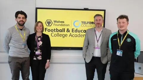 Four people stand either side of a yellow screen with the Wolves Foundation and Telford College logos on it. A man and woman are on the left of the screen, he in grey trousers and a light grey jumper, she in a black suit with a cream and black floral top. Two other men stand to the right, one in a checked blazer and white shirt and the other a Wolves black and turquoise tracksuit. All are smiling at the camera.
