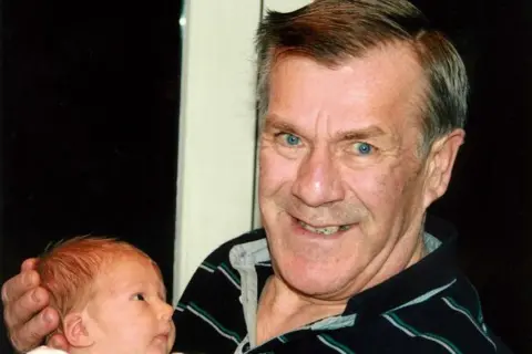 Rob and Dianne Parsons Photograph of Ronnie Lockwood(right) smiling holding a baby. He has bright blue eyes and smiles the camera. His left hand cradles the baby's head carefully.