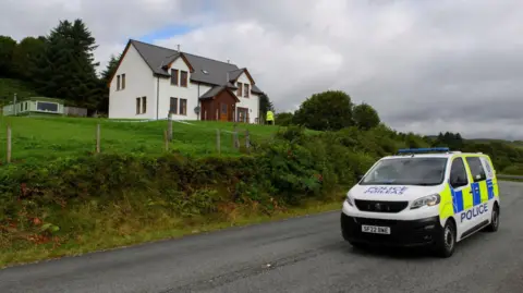 PA Media A police van is on a road below the two-storey house. There is a police officer standing in front of the property.