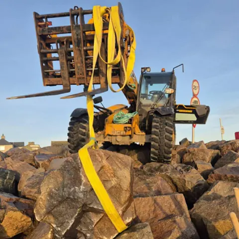 BDMLR A yellow digger-like vehicle has yellow canvas straps attached to its front loader and a large rock.