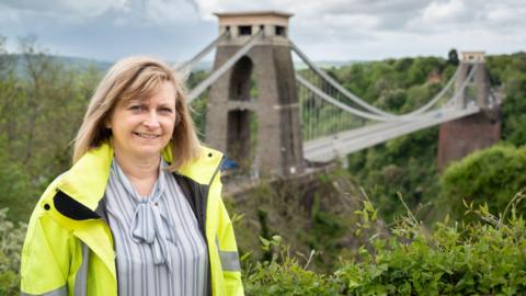 Trish Johnson standing in front of the Clifton Suspension Bridge. She is wearing a high vis jacket