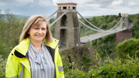 Trish Johnson Trish Johnson standing in front of the Clifton Suspension Bridge. She is wearing a high vis jacket