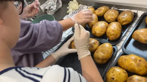 John Devine/BBC Two people preparing potatoes to be cooked as jacket spuds. One person has a fork to prick the skin, the potatoes are glossy as they have been rolled in olive oil.