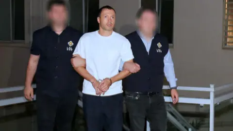 A man in a white T-shirt is held by the arms by two men in dark-coloured uniform