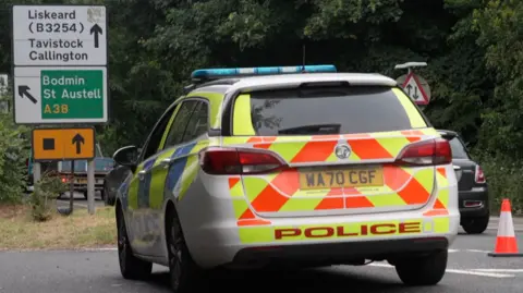 A police car is parked on a road. There is a road sign that reads: Liskeard (B3254) Tavistock Callington with an arrow indicating straight ahead. Under that it reads Bodmin St Austell A38 and points to the left. Under that sign is another sign that indicates a diversion route via a black box on orange background.