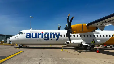 Side view of an Aurigny ATR aircraft on an airport apron under a clear blue sky, with yellow engine and black propellers visible.