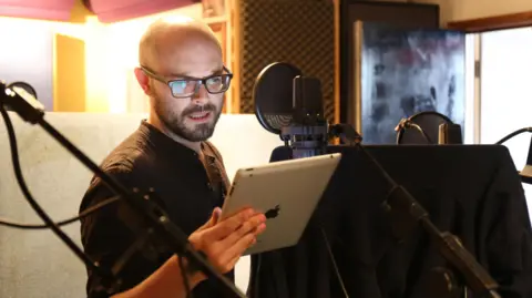Peter stands talking in front of a microphone. He is reading from a script on his iPad whilst wearing a black t-shirt. He is bald.
